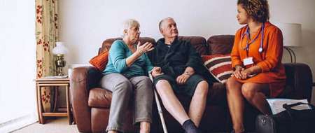 Older couple sitting on sofa talking to nurse